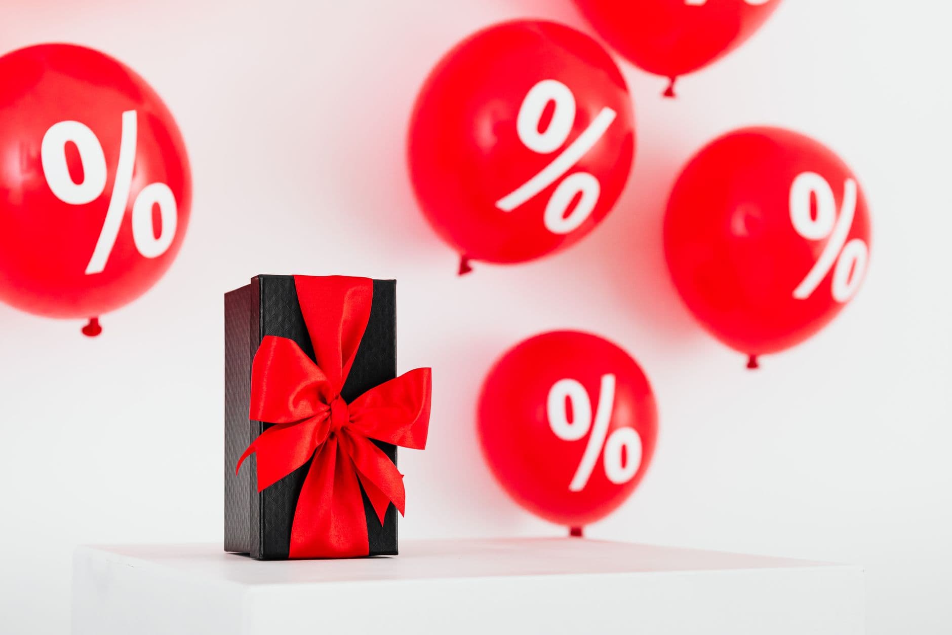 Stylish black gift box with red ribbon surrounded by red percentage balloons on a white background.