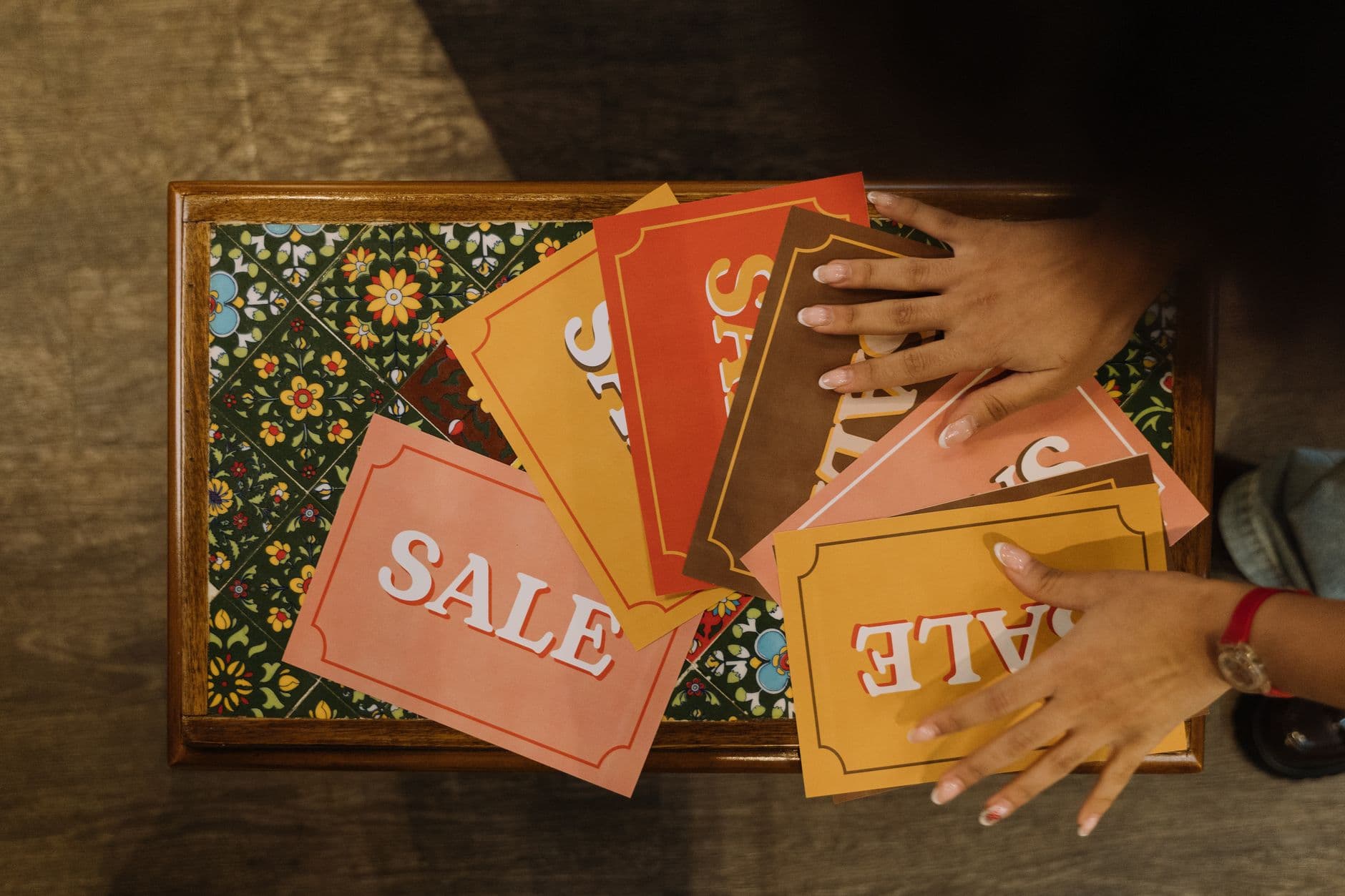 Vibrant sale signs arranged by hands on a decorative floral table surface, viewed from above.